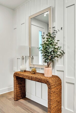 A white paneled wall with a mirror, a woven console table, a potted plant, and a white vase.