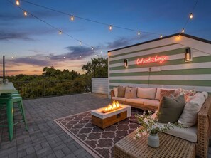 Una terraza en la azotea con un brasero, mobiliario exterior e iluminación colgante al atardecer.