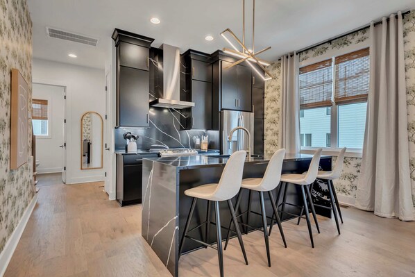 A modern kitchen with dark cabinetry, a central island with bar stools, and a large window with blinds.