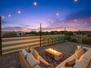 An outdoor seating area with a fire feature, string lights, and a view of the city skyline.