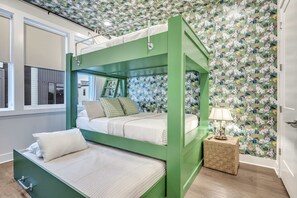 A bunk bed with a green frame, a window with blinds, a patterned wallpaper, and a small side table with a lamp.