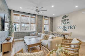 Ein gemütliches Wohnzimmer mit einer Couch, einem Couchtisch und Korbstühlen. Es gibt Fenster mit Jalousien, einen Deckenventilator und eine Wand mit einem motivierenden Zitat.