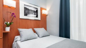 Habitación de hotel con cabecera de madera, un retrato enmarcado, dos almohadas estampadas, un jarrón con flores y una cama con manta gris.