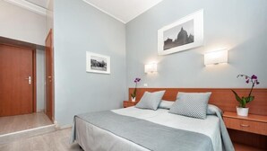 Un dormitorio con cama, mesitas de noche, cabecera de madera, luces fijadas en la pared, un cuadro enmarcado y una puerta.