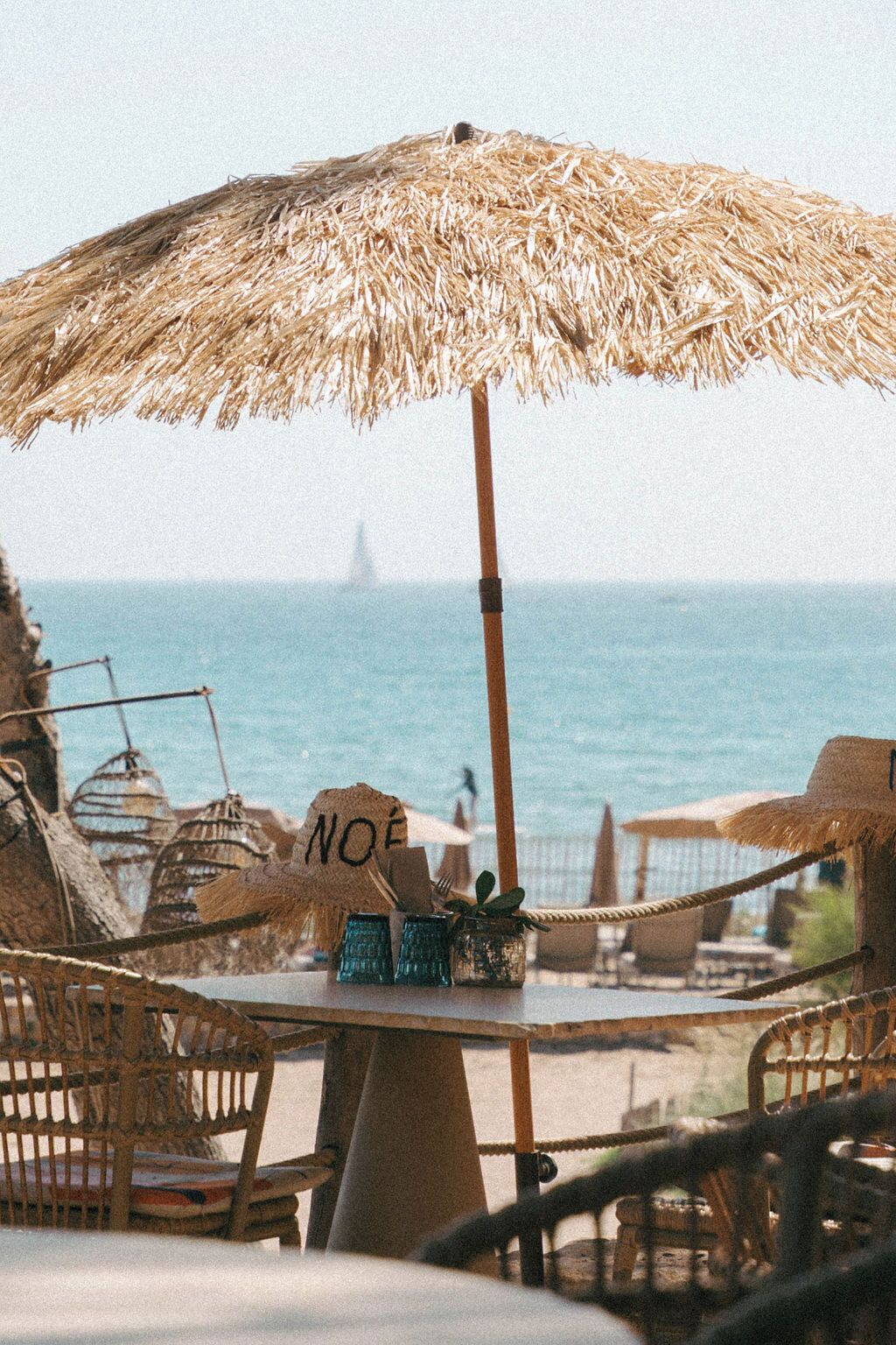 Una mesa en la playa con un paraguas de paja, sillas de mimbre y un letrero que dice 'NOÉ'.