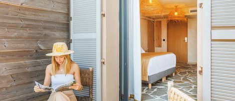 A person reading a magazine on a bench outside a room with a bed and a hanging light fixture.