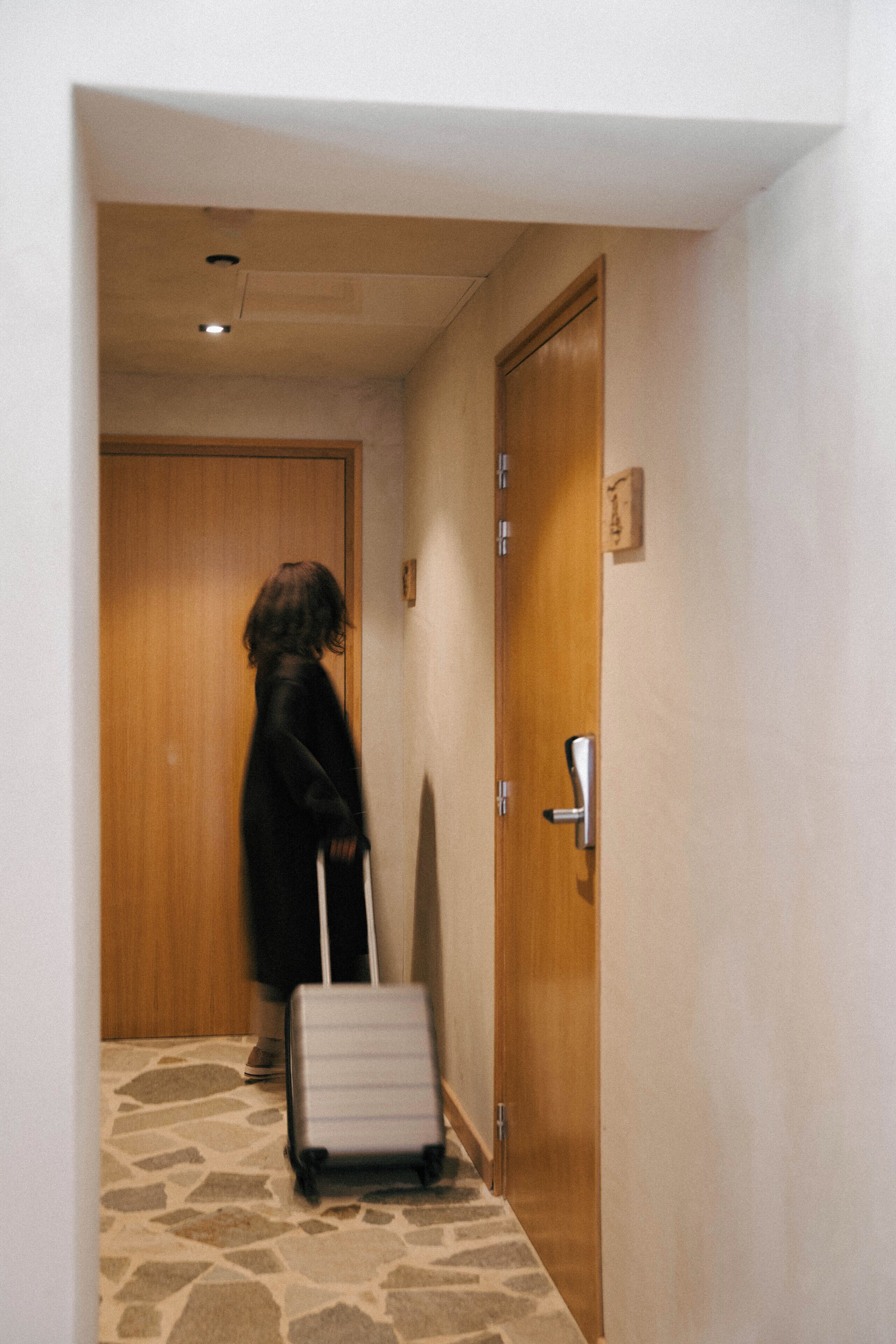 A person with a suitcase in a hallway with a wooden door and a stone floor.