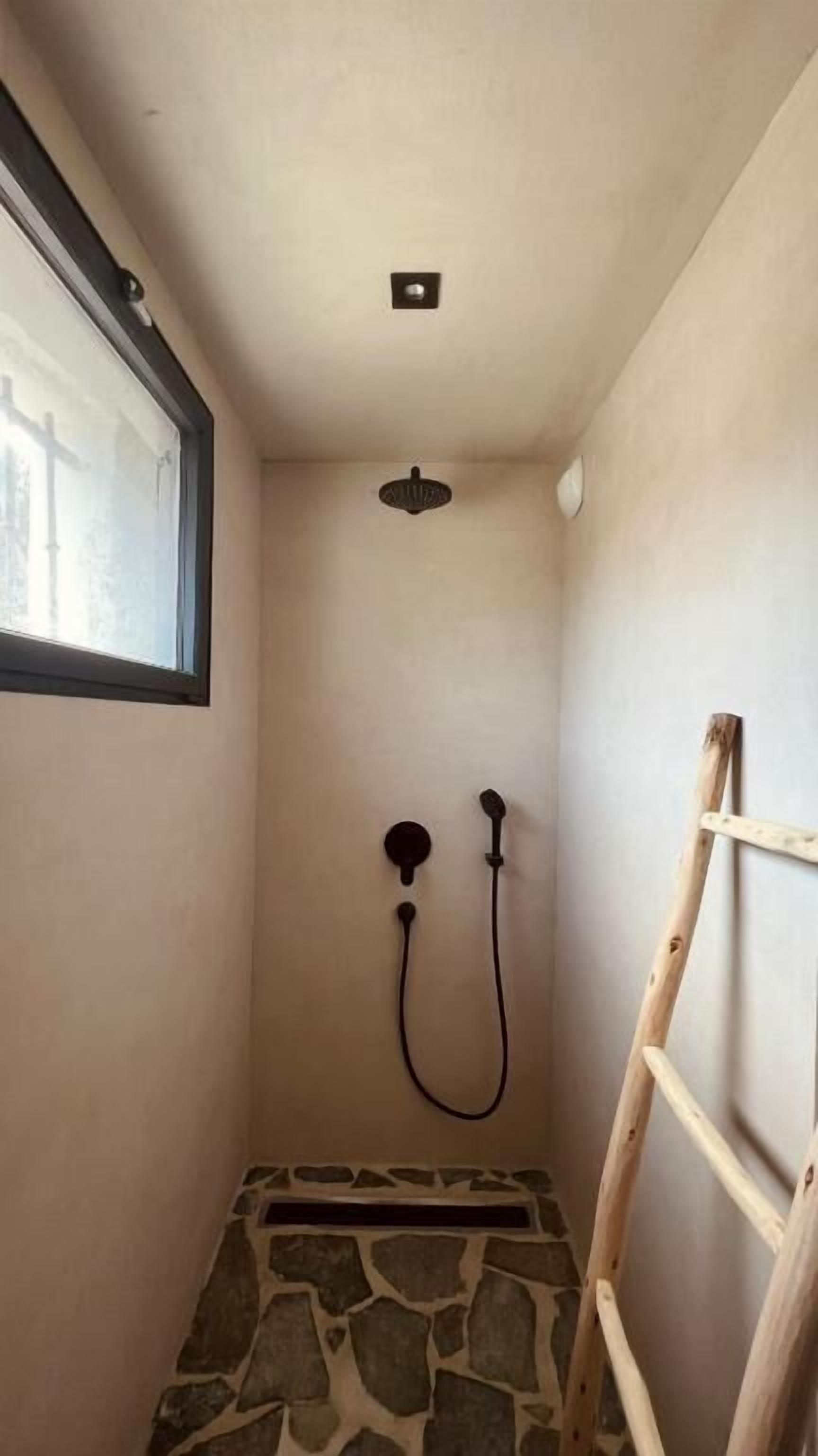 A small, stone-tiled shower room with a wooden ladder, a window, and a ceiling-mounted showerhead.