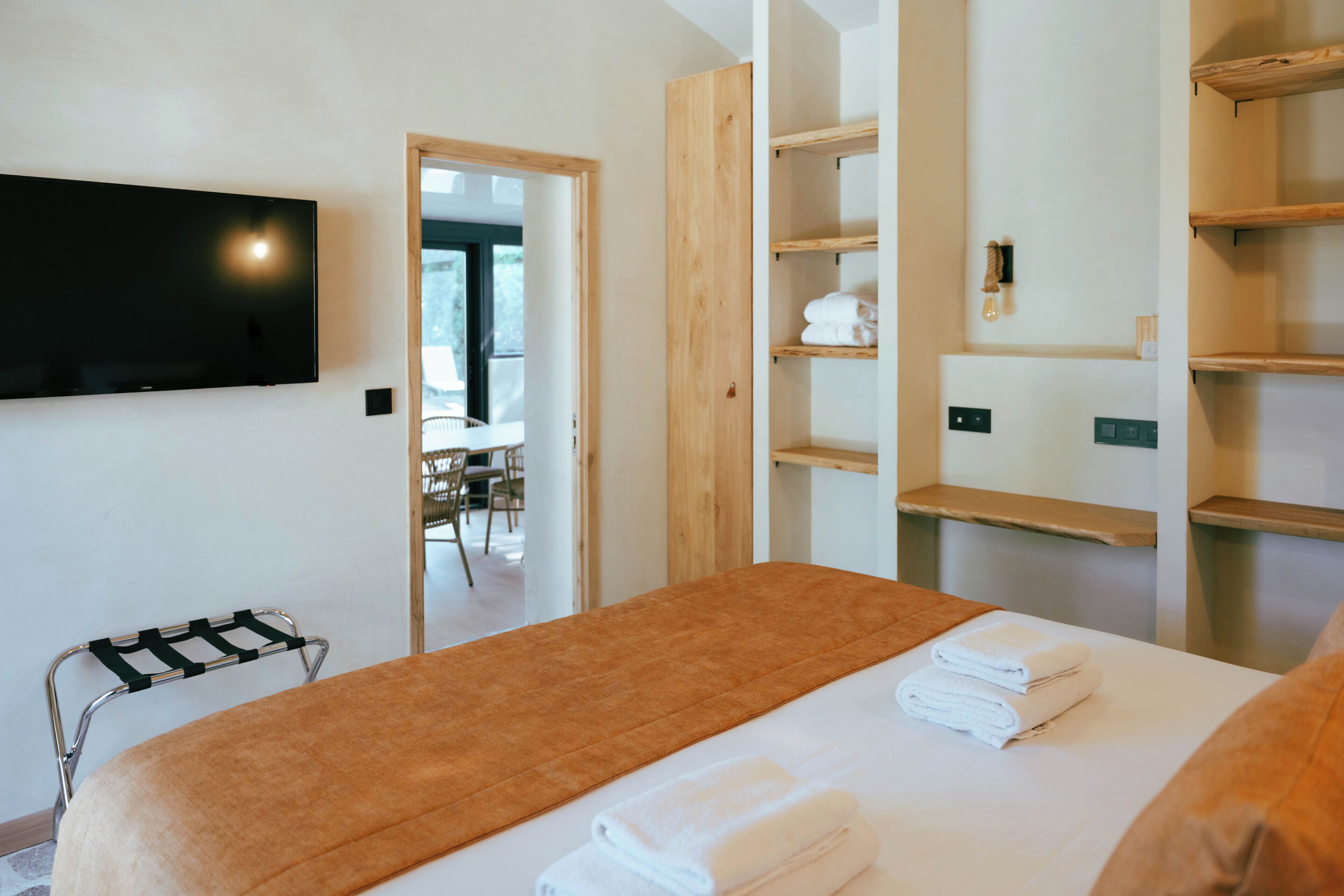 A hotel room with a bed, a TV mounted on the wall, a wooden shelf unit, and a chair with a towel rack.