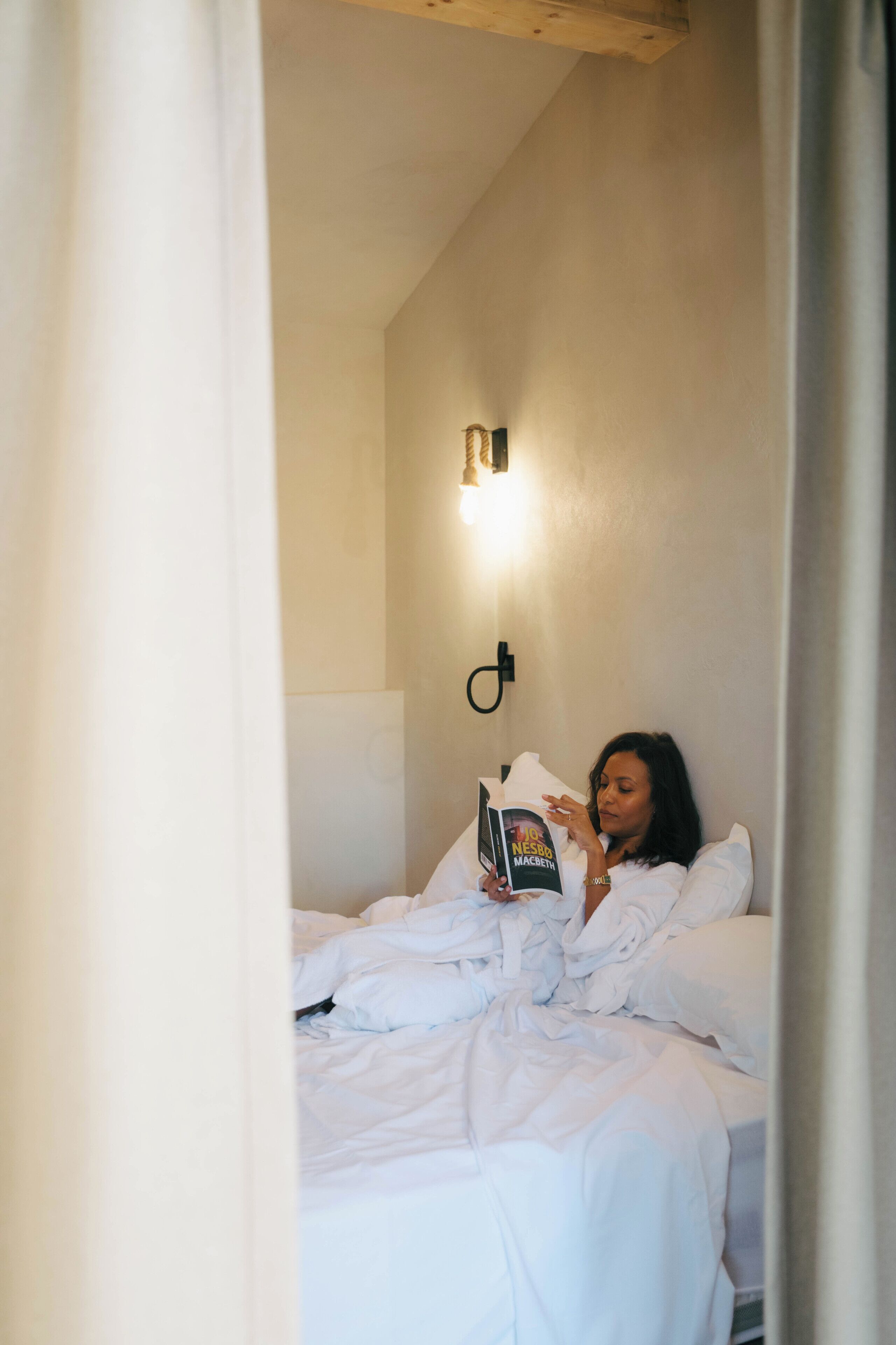 A person lying in bed reading a book in a room with a light fixture on the wall.