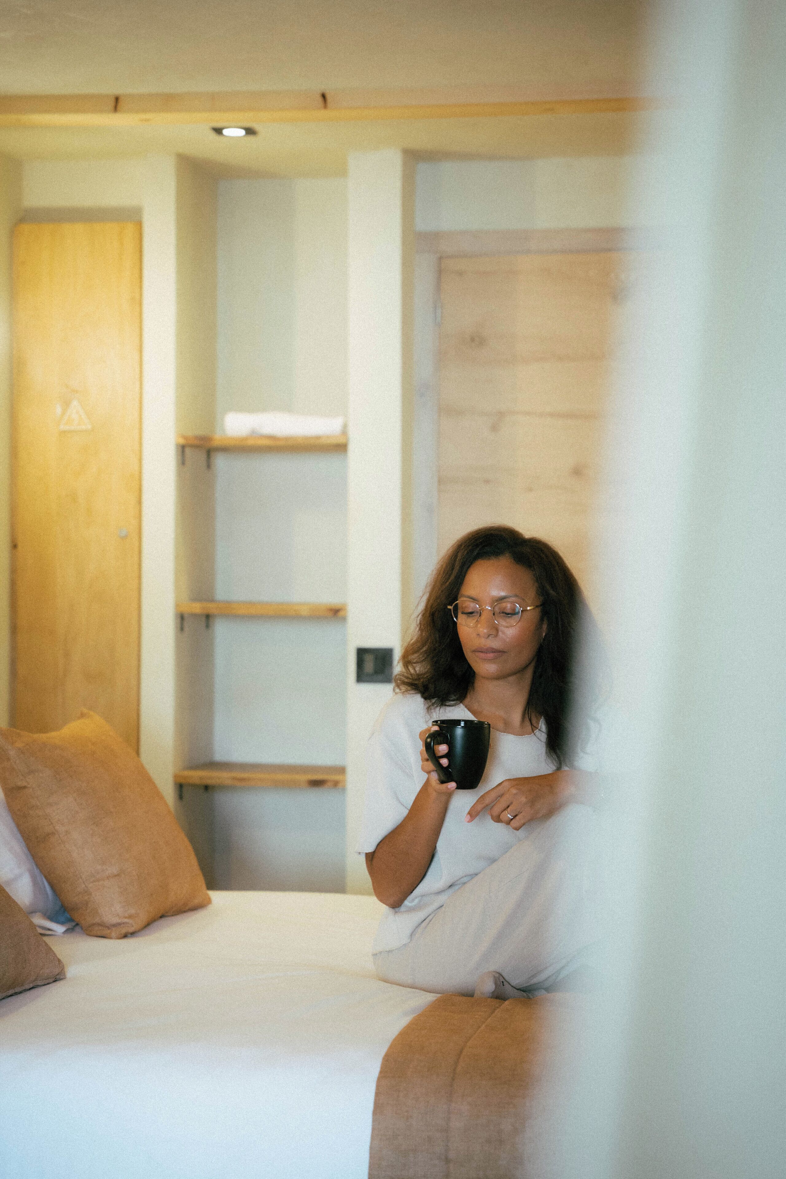 A person sitting on a bed with a black mug, in a room with wooden shelves and a door.