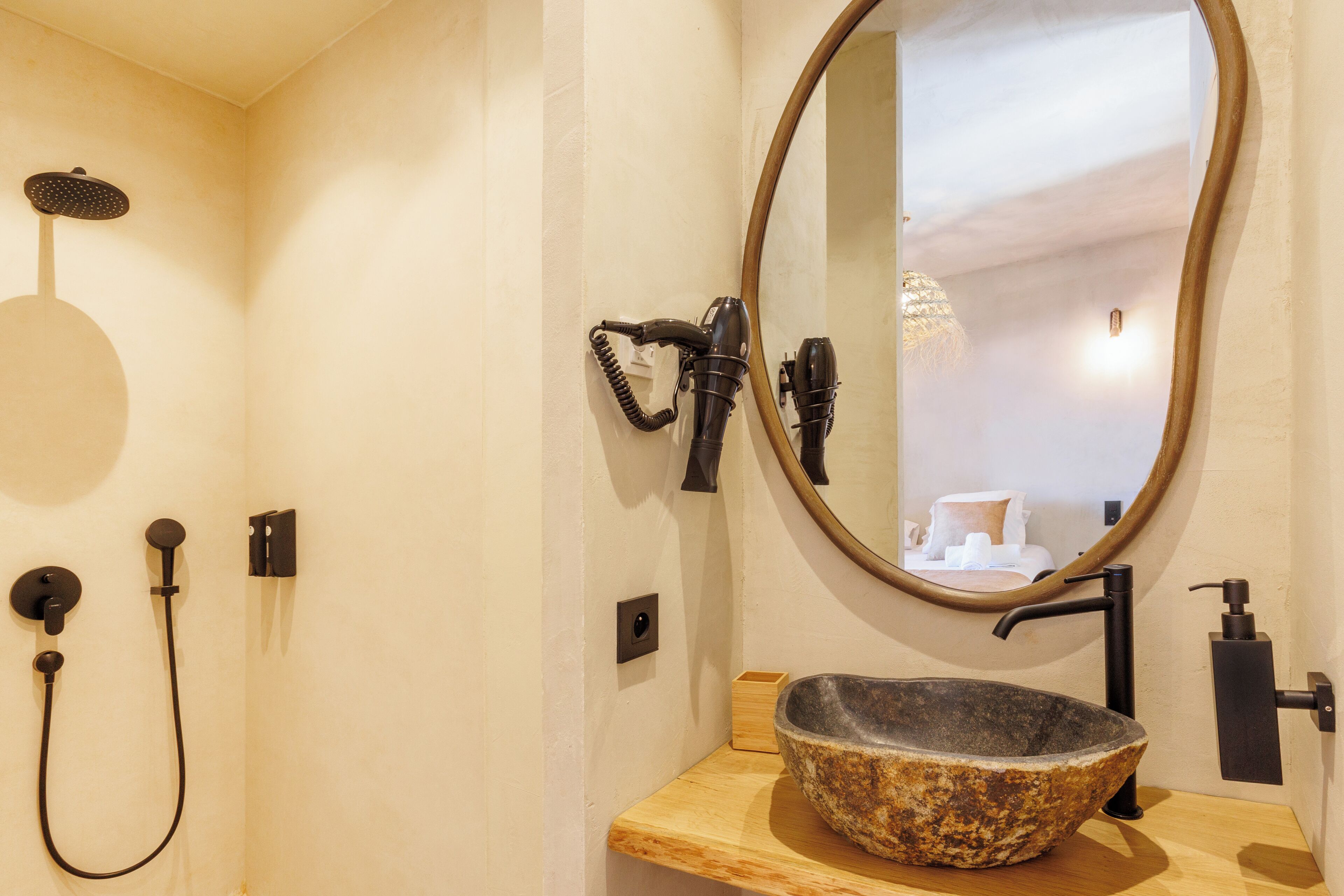 A bathroom with a stone sink, a round mirror, a handheld showerhead, and a hairdryer mounted on the wall.