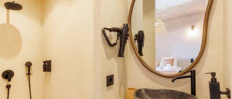 A bathroom with a stone sink, a round mirror, a handheld showerhead, and a hairdryer mounted on the wall.