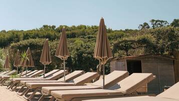 A row of sun loungers with umbrellas, a small wooden structure, and a backdrop of dense greenery.