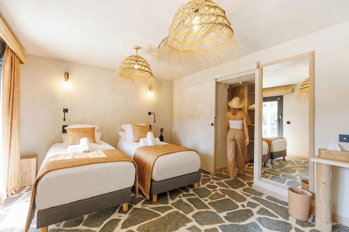 Ein Hotelzimmer mit zwei Betten, einem Boden mit Steinmuster, einem großen Spiegel und einem Holznachttisch.