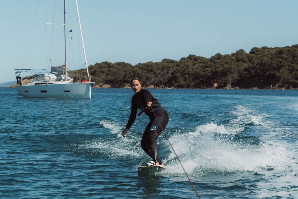 Una persona practica wakeboard sobre un cuerpo de agua, con un velero al fondo.