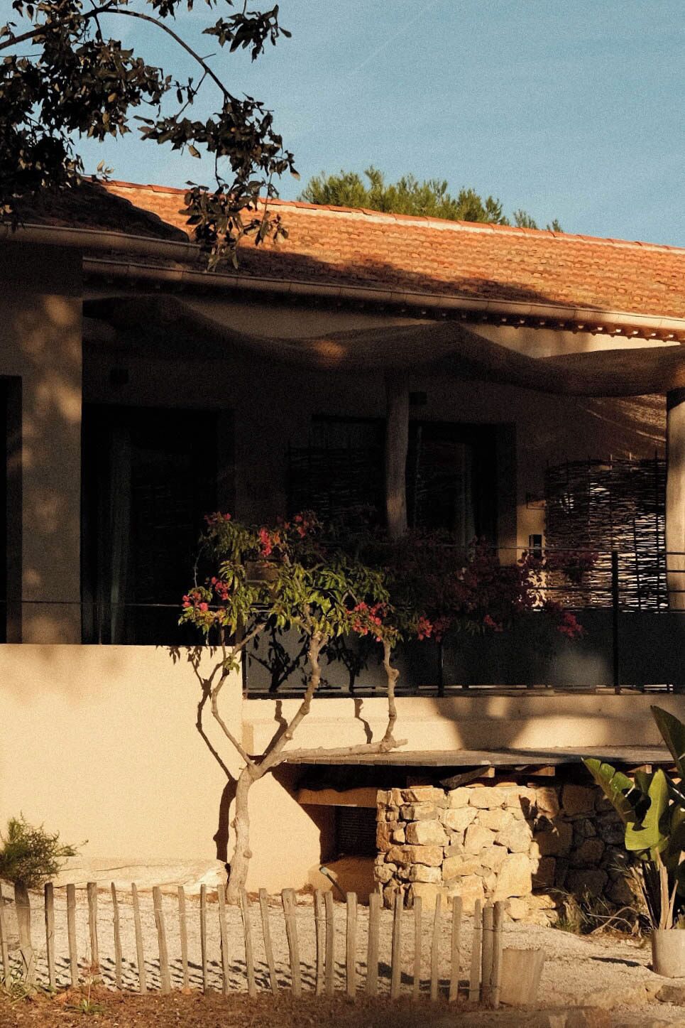 Un edificio con base de piedra, cerca de madera y un árbol con flores rosadas.