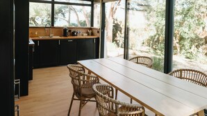 Een moderne keuken met een houten tafel en rieten stoelen, grote ramen met uitzicht op bomen, en een zwart aanrecht met een koffiezetapparaat.