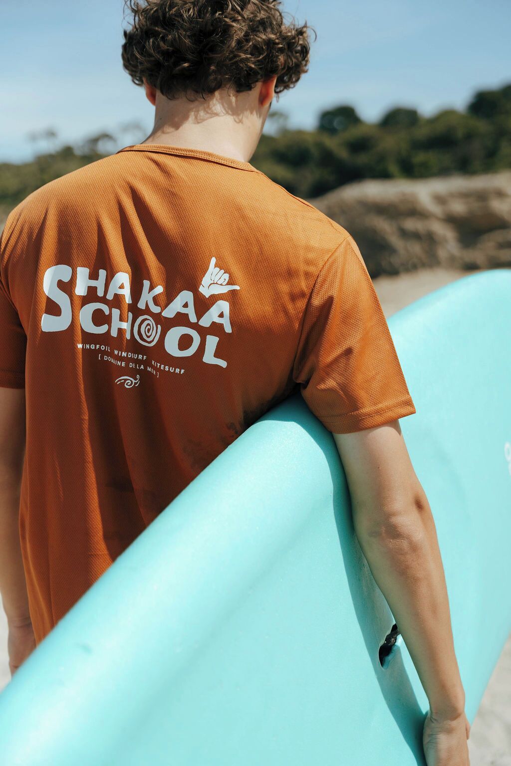 Una persona con camiseta naranja que tiene el texto "Shaka School" en la espalda, sostiene una tabla de surf azul claro.
