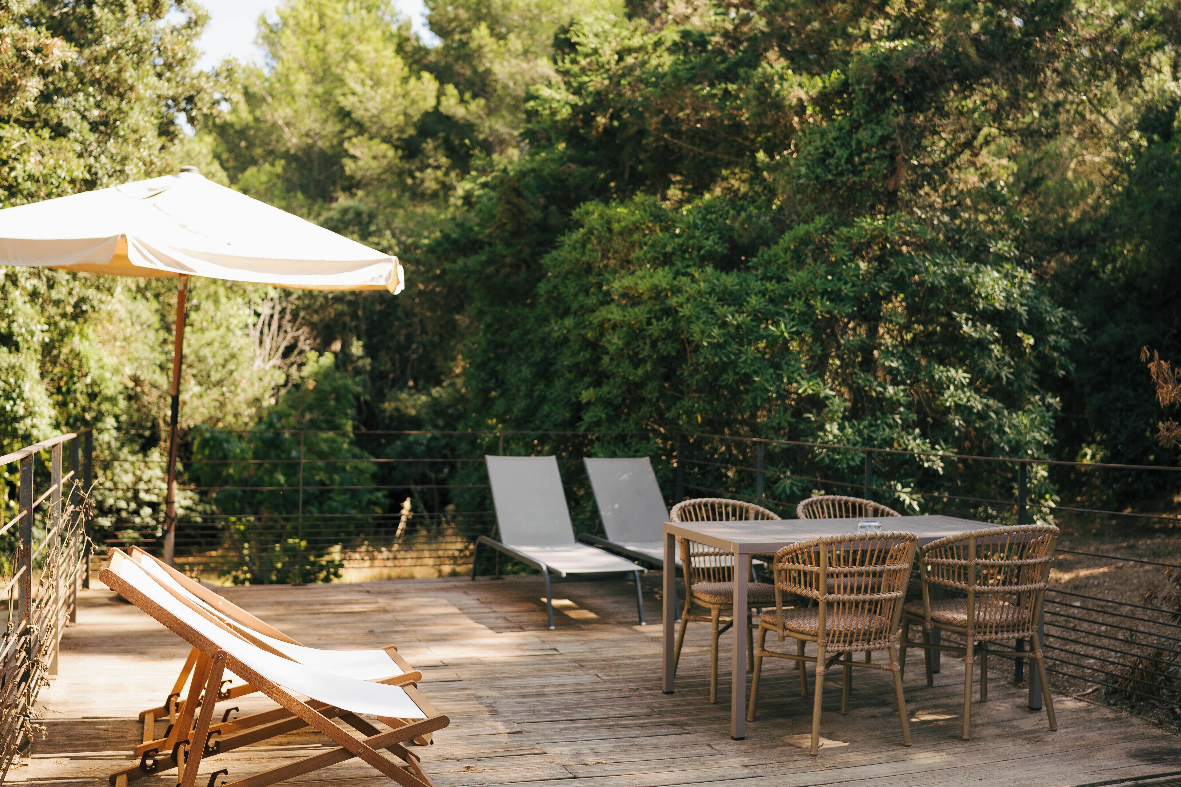 Une terrasse en bois avec des chaises longues et une table entourée de chaises, le tout bordé d’arbres.