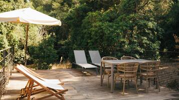 Une terrasse en bois avec des chaises longues et une table entourée de chaises, le tout bordé d’arbres.