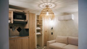 Een ruimtelijk ingerichte leefruimte met kitchenette, een geweven lamp, een beige bank en een vloer met een stenen patroon.