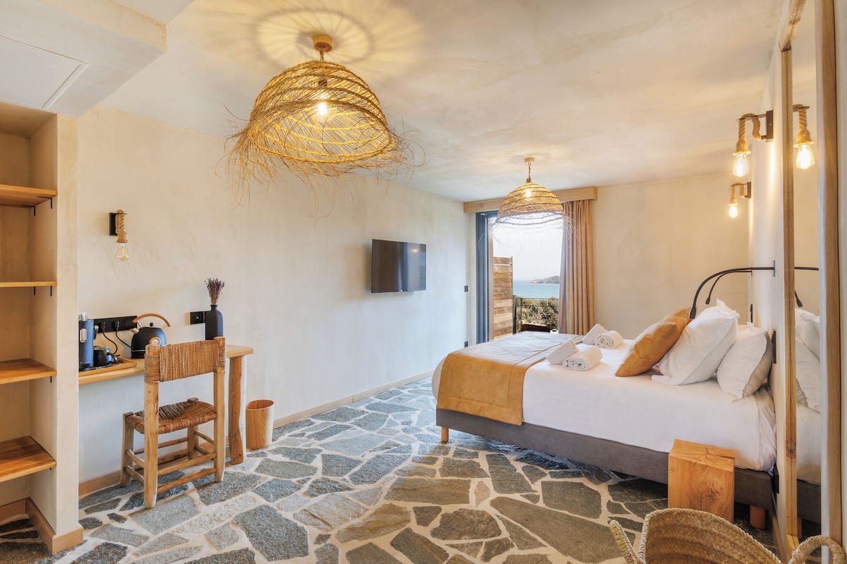 Ein Schlafzimmer mit Steinfußboden, einem Bett mit weißen Leinen, einem hölzernen Schreibtisch, einem Sessel und einem Fernseher an der Wand.