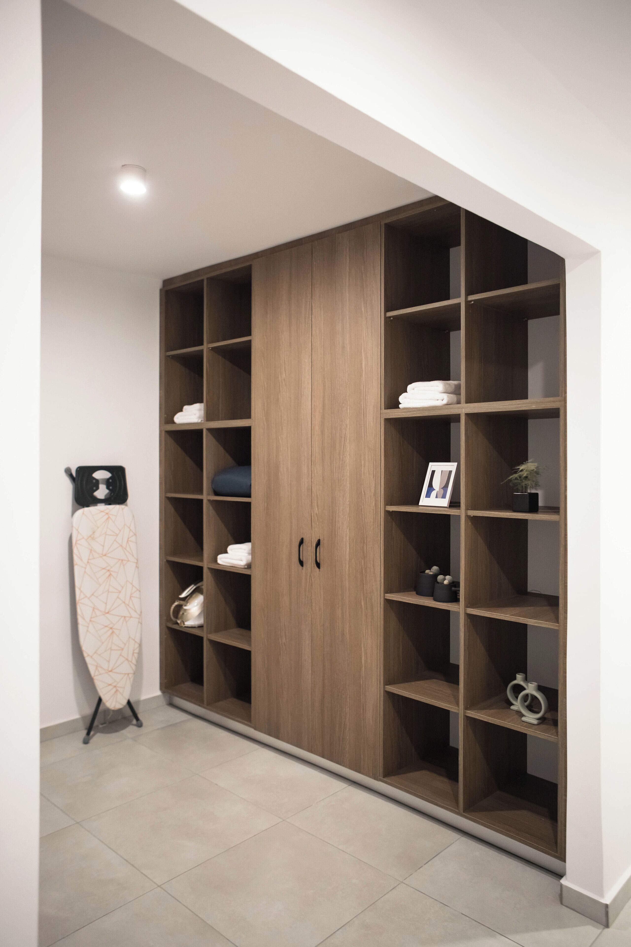 A large wooden wardrobe with multiple shelves and hanging space, a ironing board, and a small potted plant.