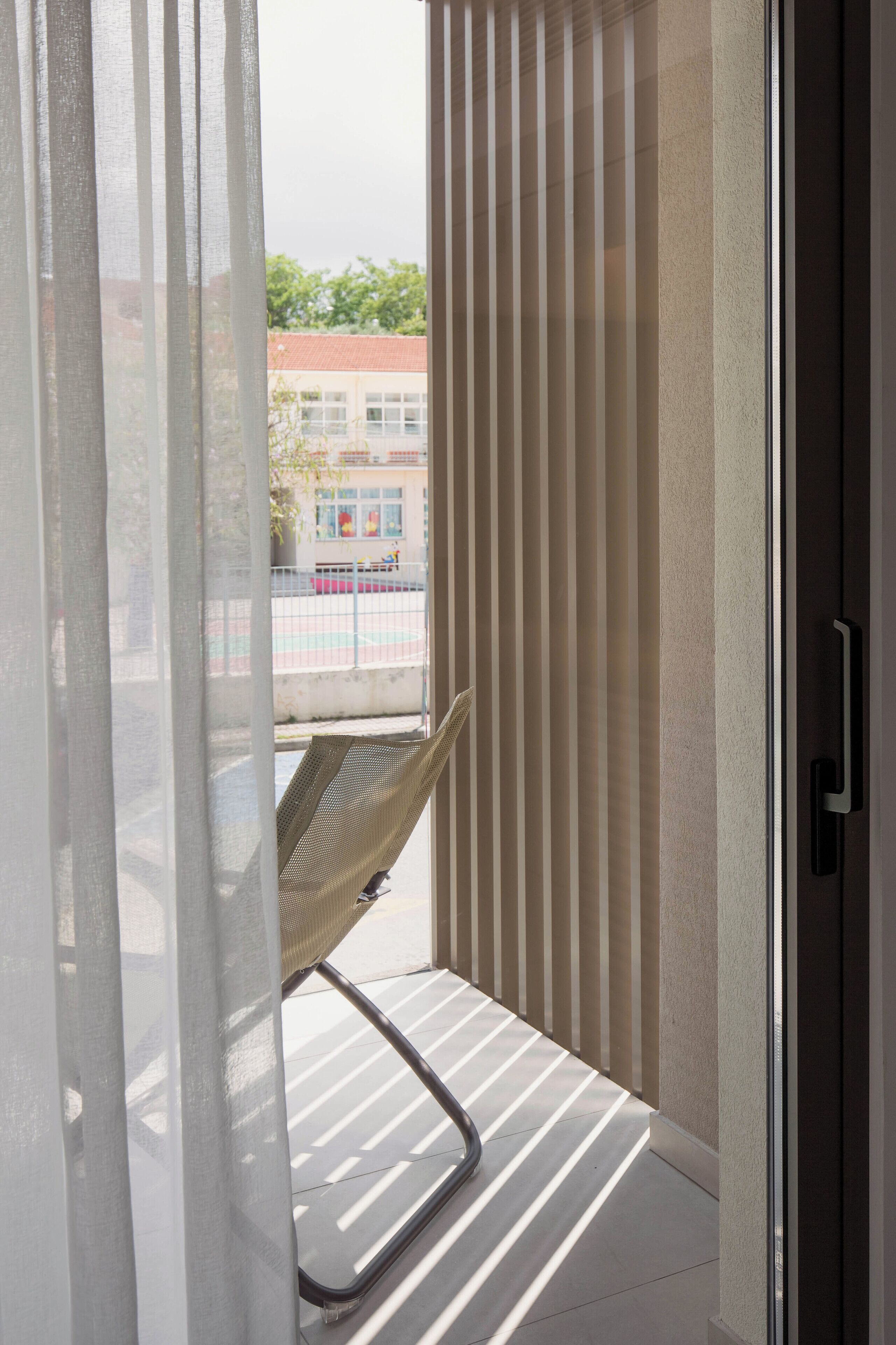 A balcony with a chair, a sliding glass door, and a view of a building with a red roof.