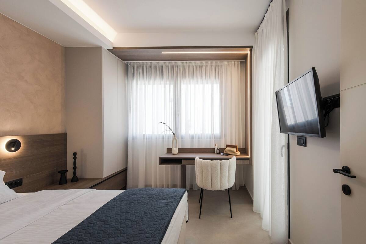 A hotel room with a bed, a desk with a chair, a TV, and a window with curtains.