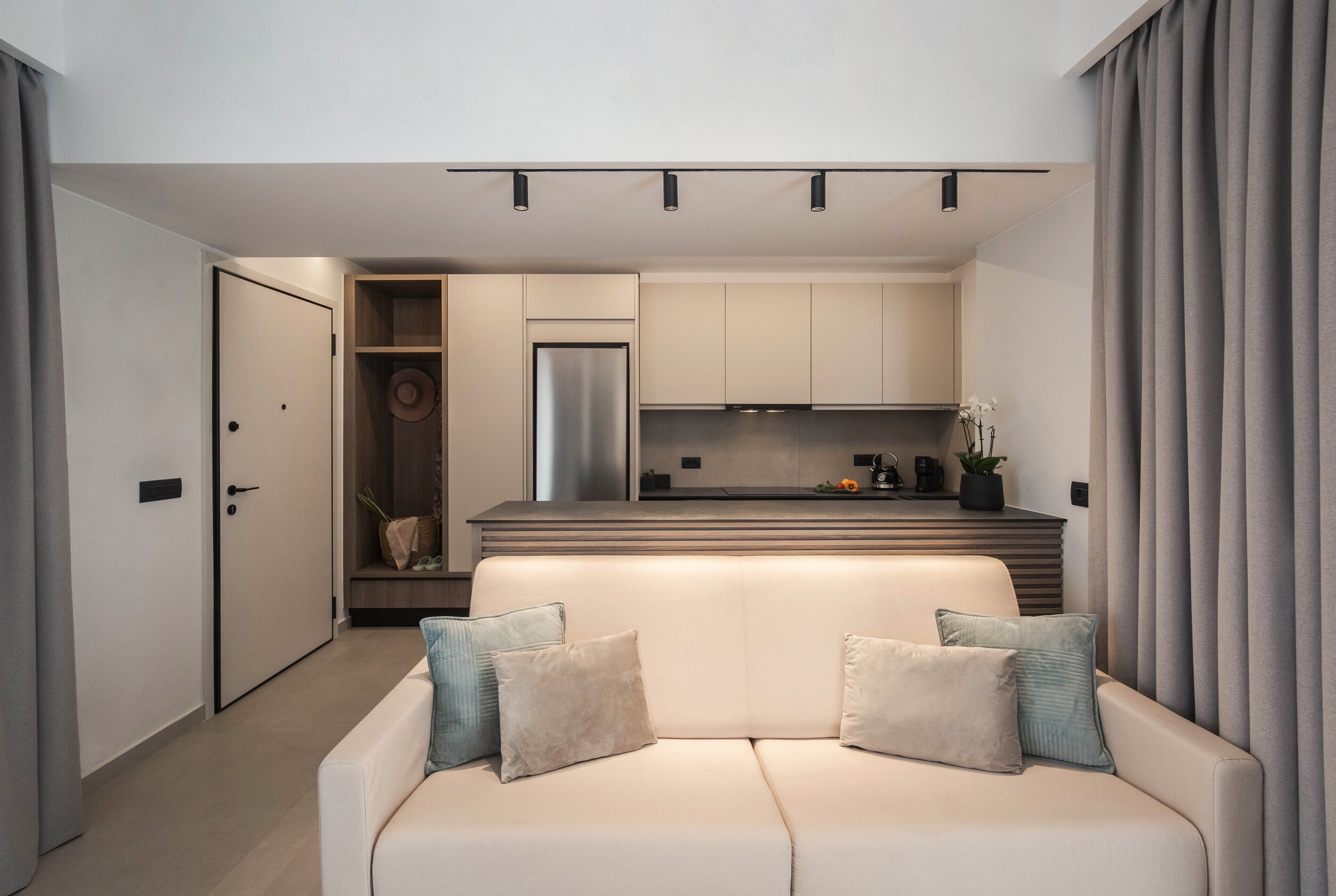 A modern living room with a beige sofa, a small kitchen area, and a door leading to another room.
