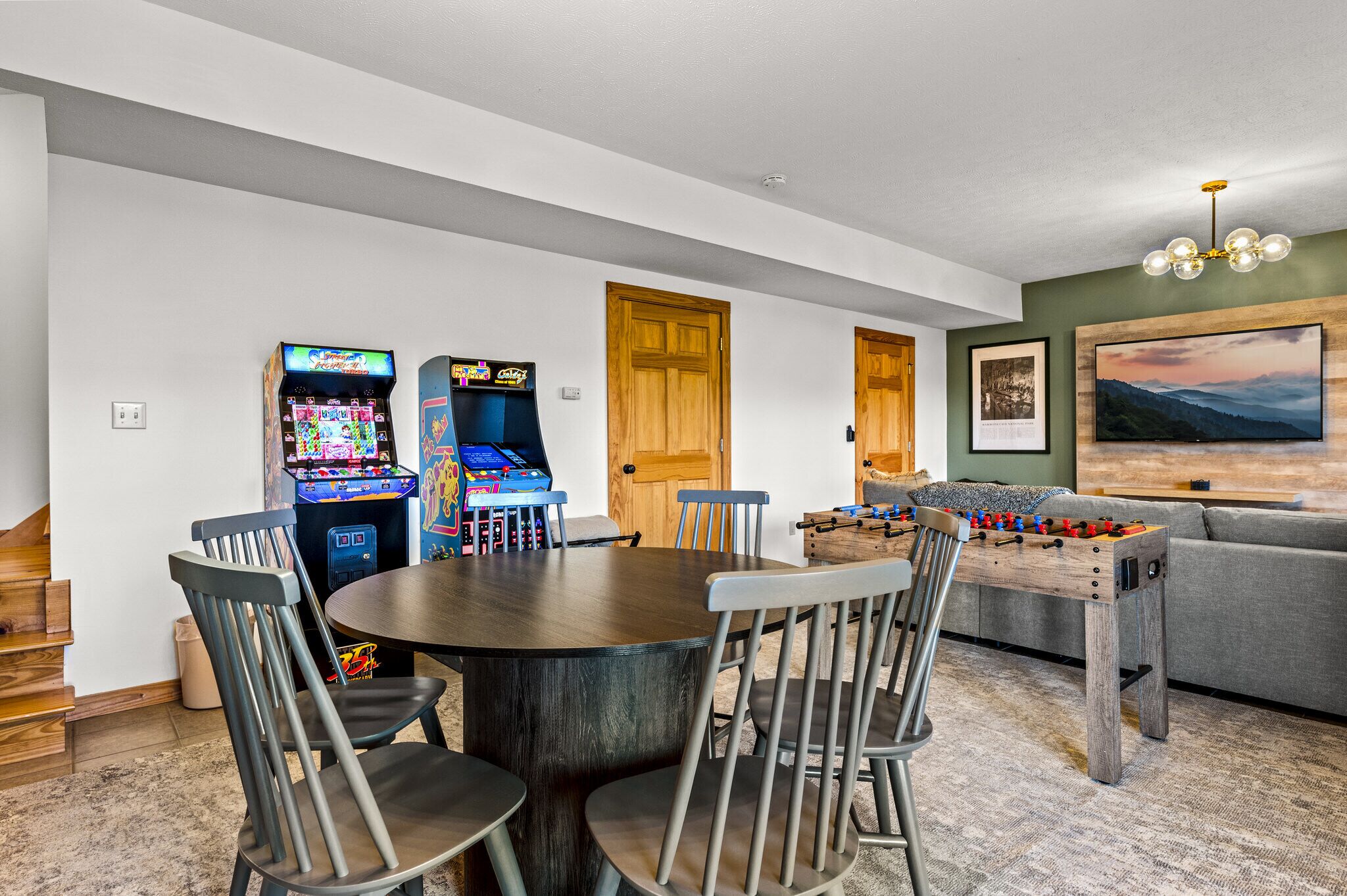 A room with a round table, four chairs, two arcade games, a foosball table, and a painting on the wall.