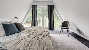 Ein Schlafzimmer mit schräg angesetzter Decke, einem Bett mit gemustertem Überwurf, einem Stuhl und Blick auf Grünfläche draußen.