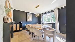 Ein moderner Essbereich mit einem Holztisch, weißen Stühlen, einer dunkel abgesetzten Wand und einem Fenster mit Vorhängen.