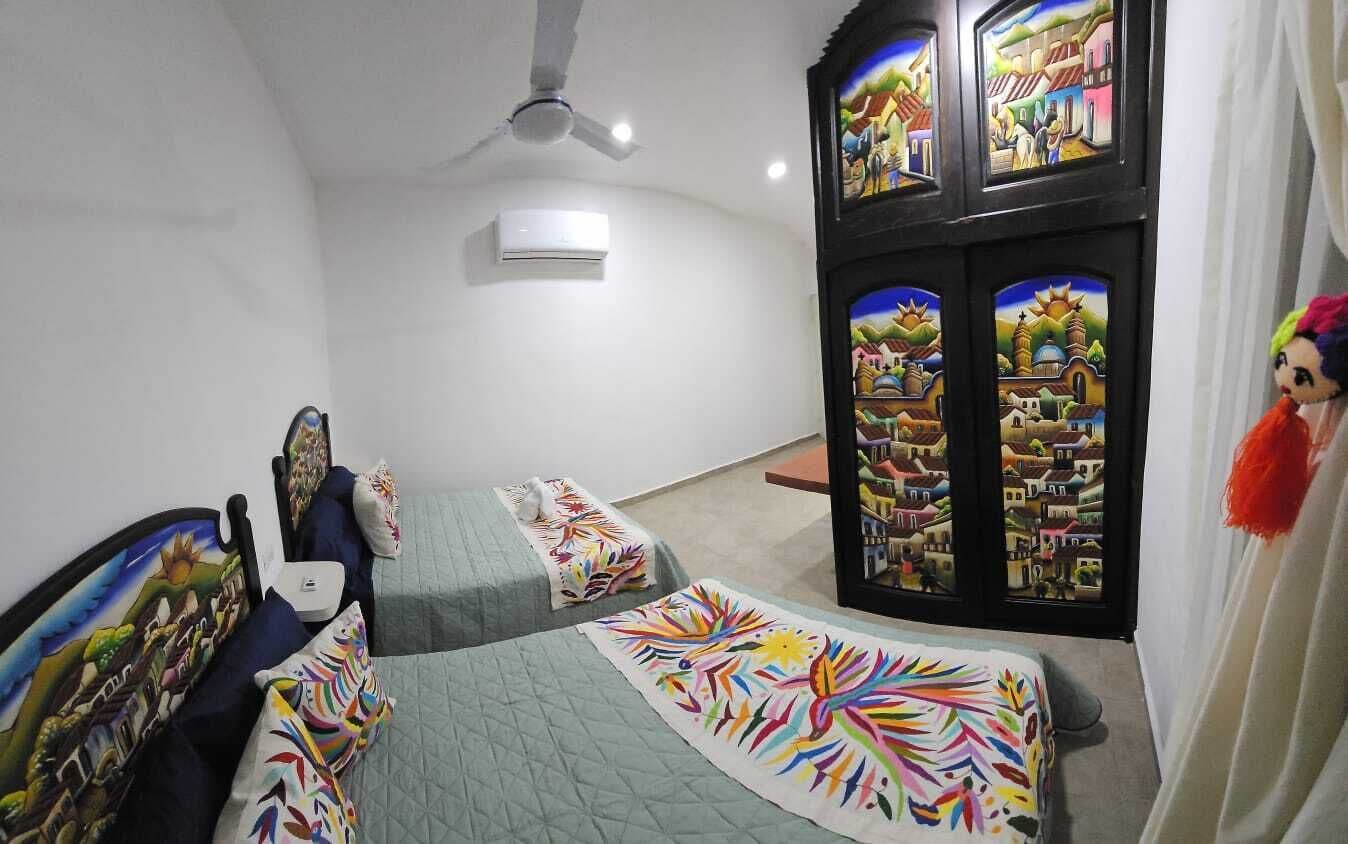 Una stanza con due letti, un ventilatore a soffitto, un condizionatore e una finestra con vista sulle case.