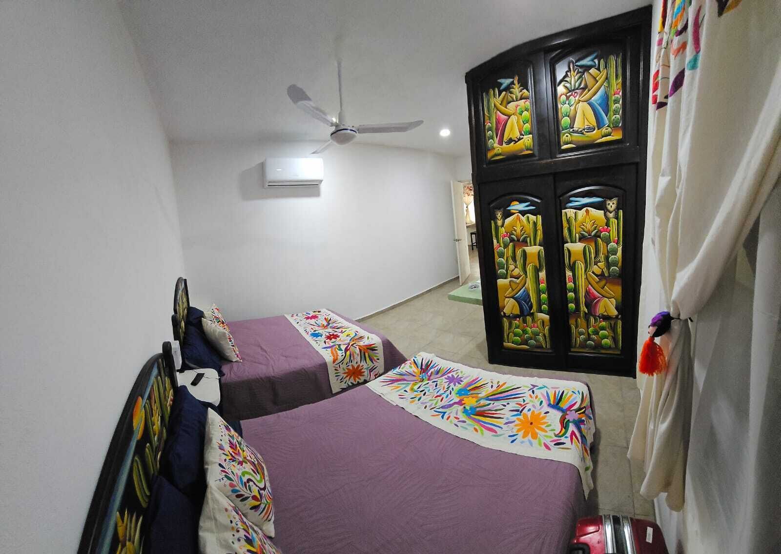 A bedroom with two beds, a ceiling fan, an air conditioning unit, and a window with colorful artwork.