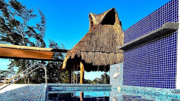 A swimming pool with blue mosaic tiles, a thatched-roof structure, and a building with blue-tiled walls. - Hotel Villas Ponto Holbox (Isla Holbox)