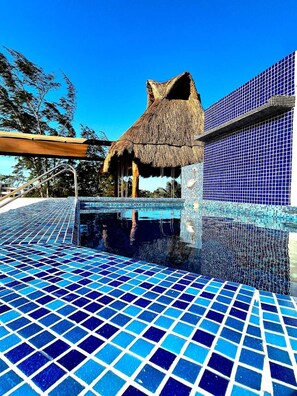 A swimming pool with blue mosaic tiles, a thatched-roof structure, and a building with blue-tiled walls. - Hotel Villas Ponto Holbox (Isla Holbox)