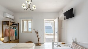 A bedroom with a bed, a bedside table, a TV, a wardrobe, and a window with a view of the sea. - Anemi Suites (Milos)