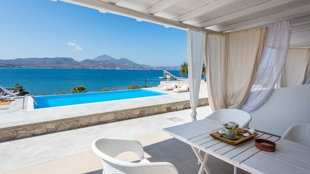 A balcony with a view of a blue sea and mountains, a white table with a tray, and a white chair. - Anemi Suites (Milos)