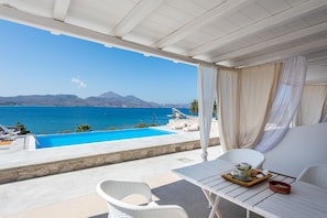 A balcony with a view of a blue sea and mountains, a white table with a tray, and a white chair.