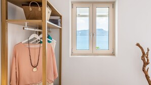 A room with a wardrobe containing clothes and a hanging bag, a window with a view of the sea, and a wooden branch decoration. - Anemi Suites (Milos)