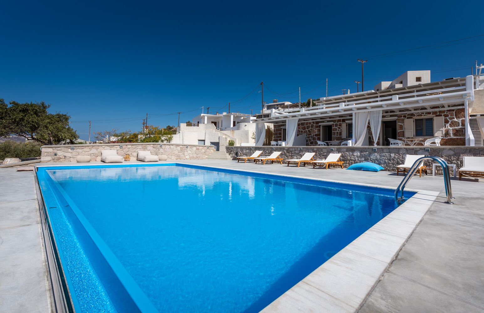 Una piscina rectangular con agua azul cristalina, rodeada de sillas reclinables blancas y un muro de piedra.