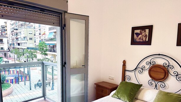 Un dormitorio con cama, cabecera de madera, almohada verde, mesita de noche de madera y vista a un balcón con paisaje urbano.