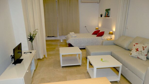 Ein modernes Wohnzimmer mit einer Couch, zwei weißen Couchtischen, einem Fernseher auf einem Schrank und einem Bett mit roten Kissen.