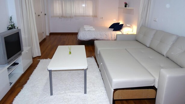 A modern, well-lit room with a white sofa, a small table, a TV, and a bed with a nightstand. - Minimalist center loft (Córdoba)