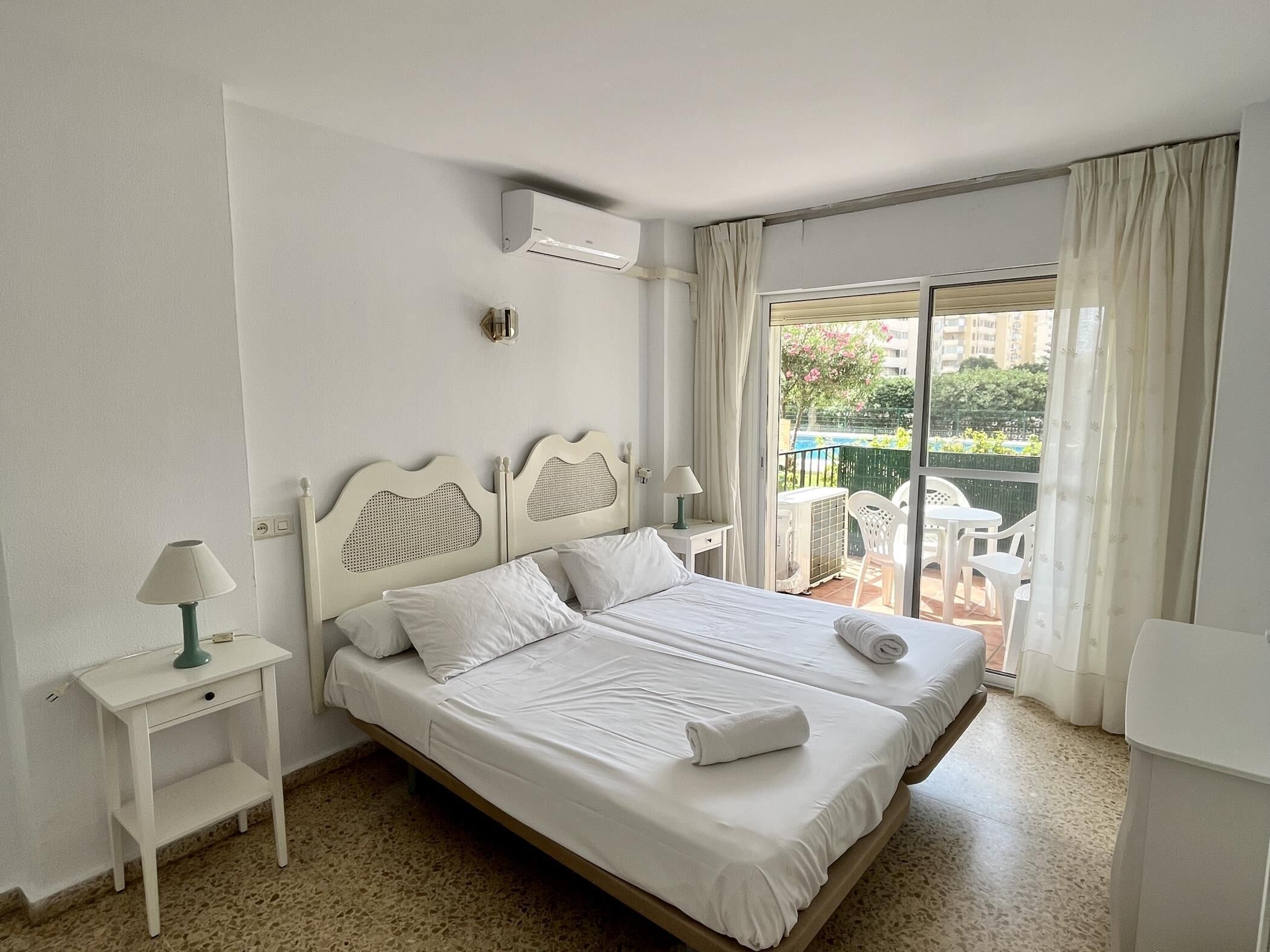 A hotel room with a bed, a nightstand, a lamp, a window with curtains, and a view of the outside balcony.