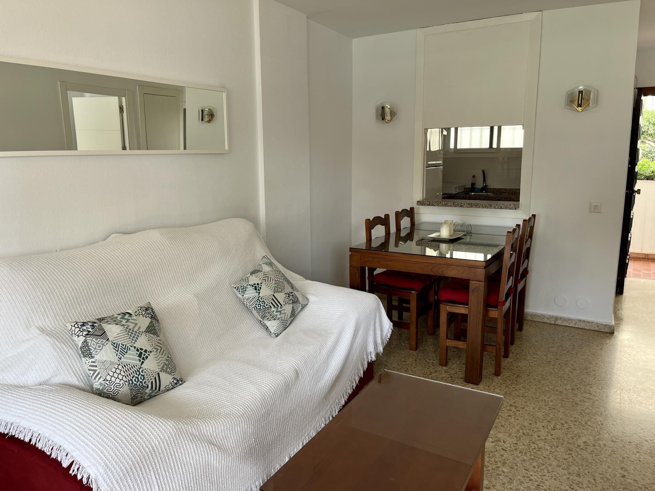 Una habitación con sofá blanco, almohadones estampados, mesa de comedor con superficie de vidrio, sillas de madera, zona de cocina con mostrador y armarios, y una puerta que da al exterior.