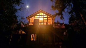 Une cabane en bois dotée d’une grande fenêtre, entourée d’arbres, et une lune dans le ciel.