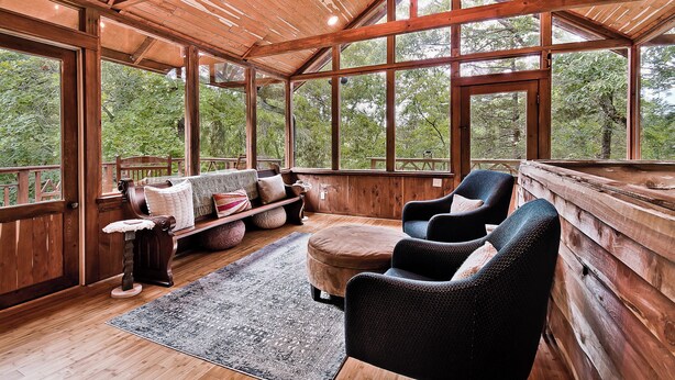 Une cabane en bois dotée de grandes fenêtres, d’une terrasse en bois, d’un canapé et de deux fauteuils.